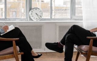 Person in black pants and black shoes sitting on brown wooden chair
